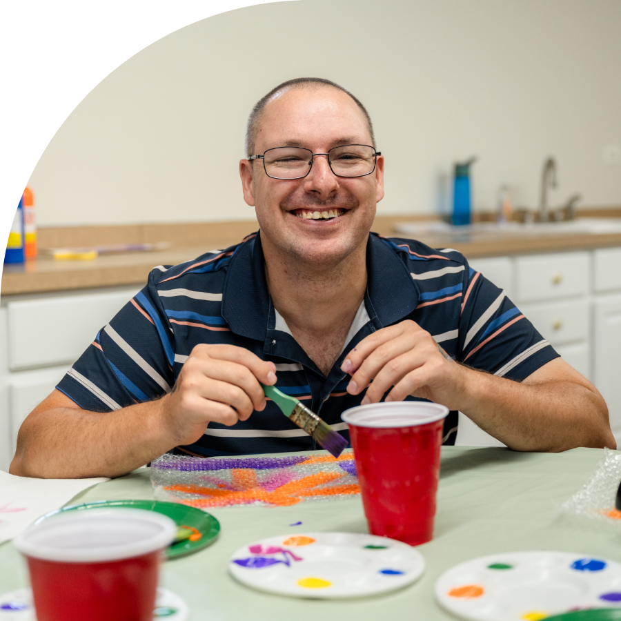 A man in a blue shirt and glasses paints a colorful craft project while smiling at the camera.